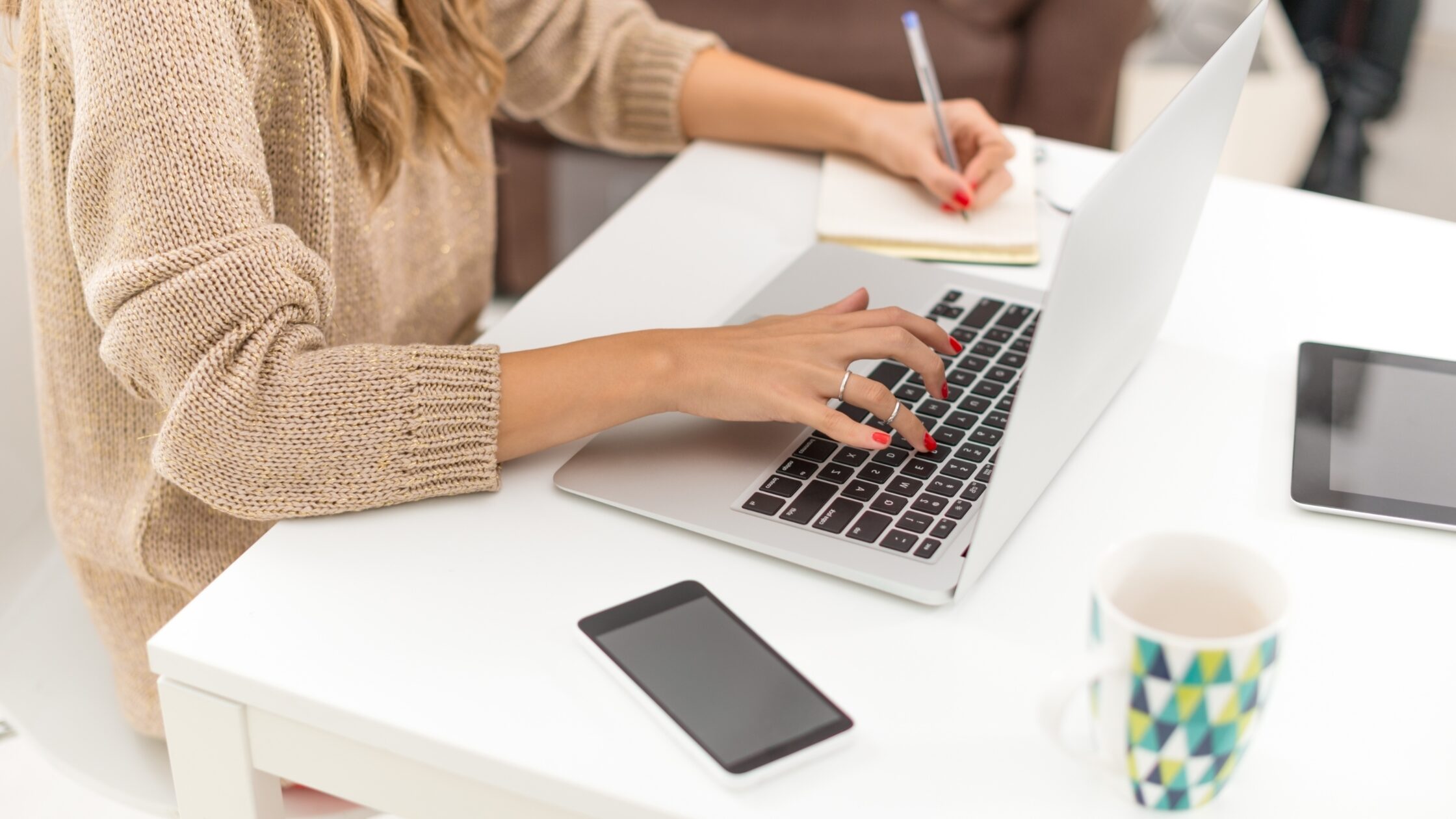 Close-up shot of woman's hands, typing on a laptop keyboard, while taking notes in a notebook
