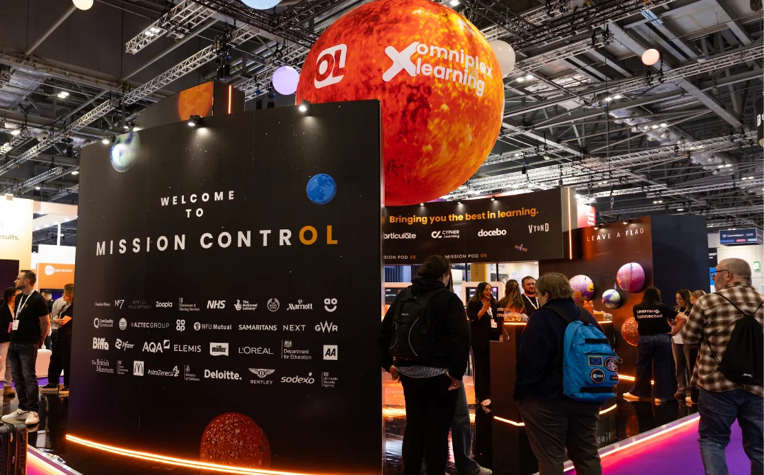 A photo of the Omniplex Learning stand at Learning Technologies - featuring a big orange sun hovering from the ceiling and people visiting the stand.