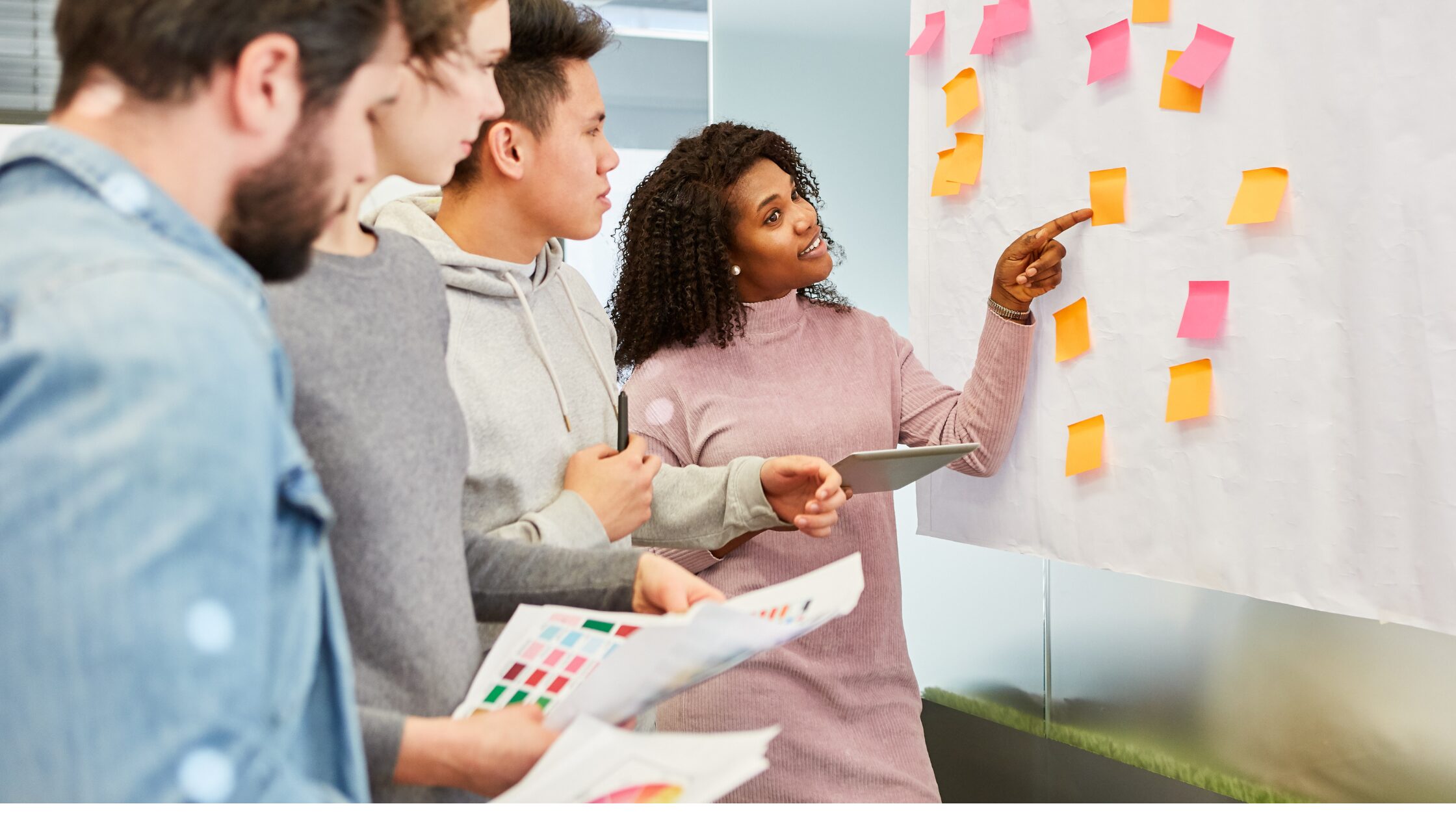 Group of colleagues gathered around a whiteboard during a planning meeting, with the leader pointing to sticky notes on the board