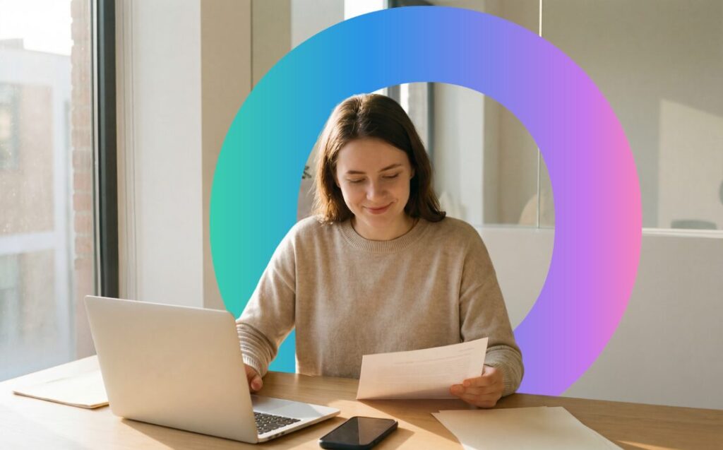 Woman sat a desk doing admin; she's typing on a laptop with one hand and holding a piece of paper with the other