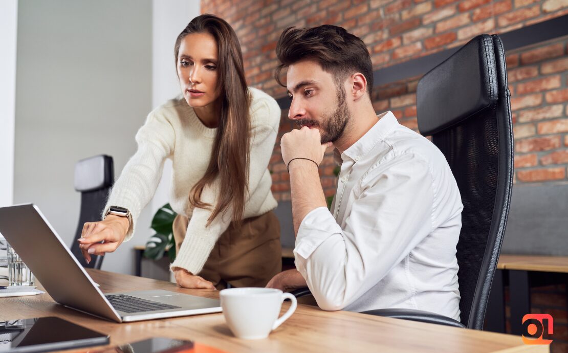 Man looking at laptop thoughtfully, while a woman stood next to him points at the screen, instructing him how to do a task