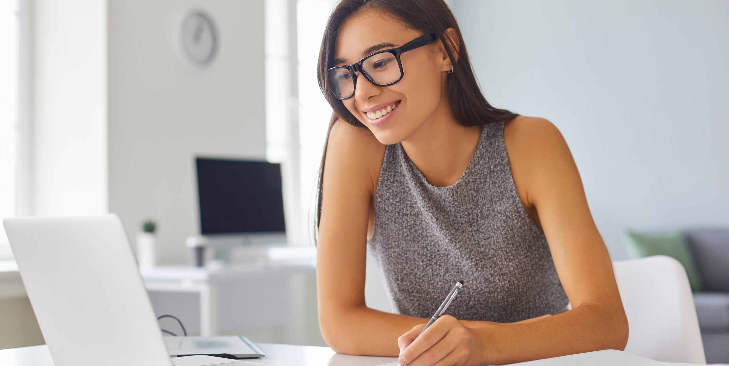 Woman smiling at laptop, while holding a pen to take notes