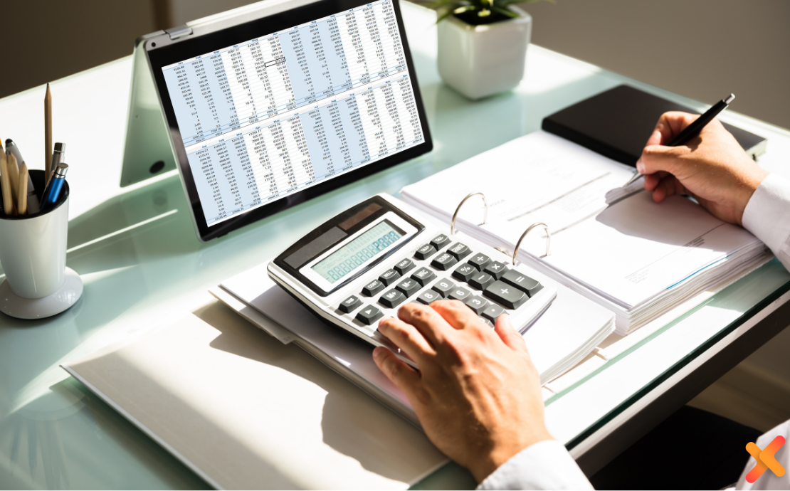 Close up shot of a desk, showing a tablet with a spreadsheet of numbers. A man's hands are on the desk too; one hand on a calculator, and the other holding a pen