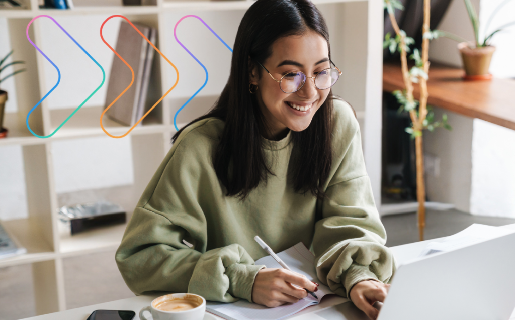 A young woman with glasses smiles while working on a laptop and taking notes in a notebook at a desk, with a coffee cup nearby