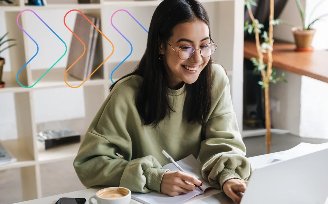 A young woman with glasses smiles while working on a laptop and taking notes in a notebook at a desk, with a coffee cup nearby